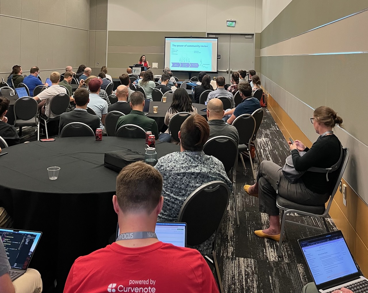 Image of a room of round tables with black covers filled with people listening to my talk. At the front is a speaker wearing a red tank top. On the screen is a slide that says 'The Power of Community Review.'