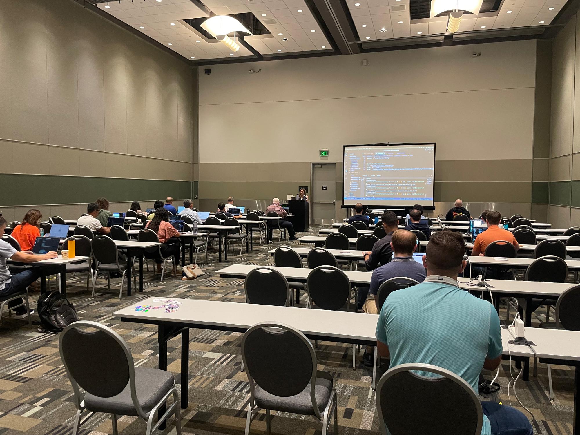 A photograph showing a large open and well-lit meeting room with two rows of horizontal tables. People are working at laptops in the room. An instructor is at the front with their laptop screen projected on the screen