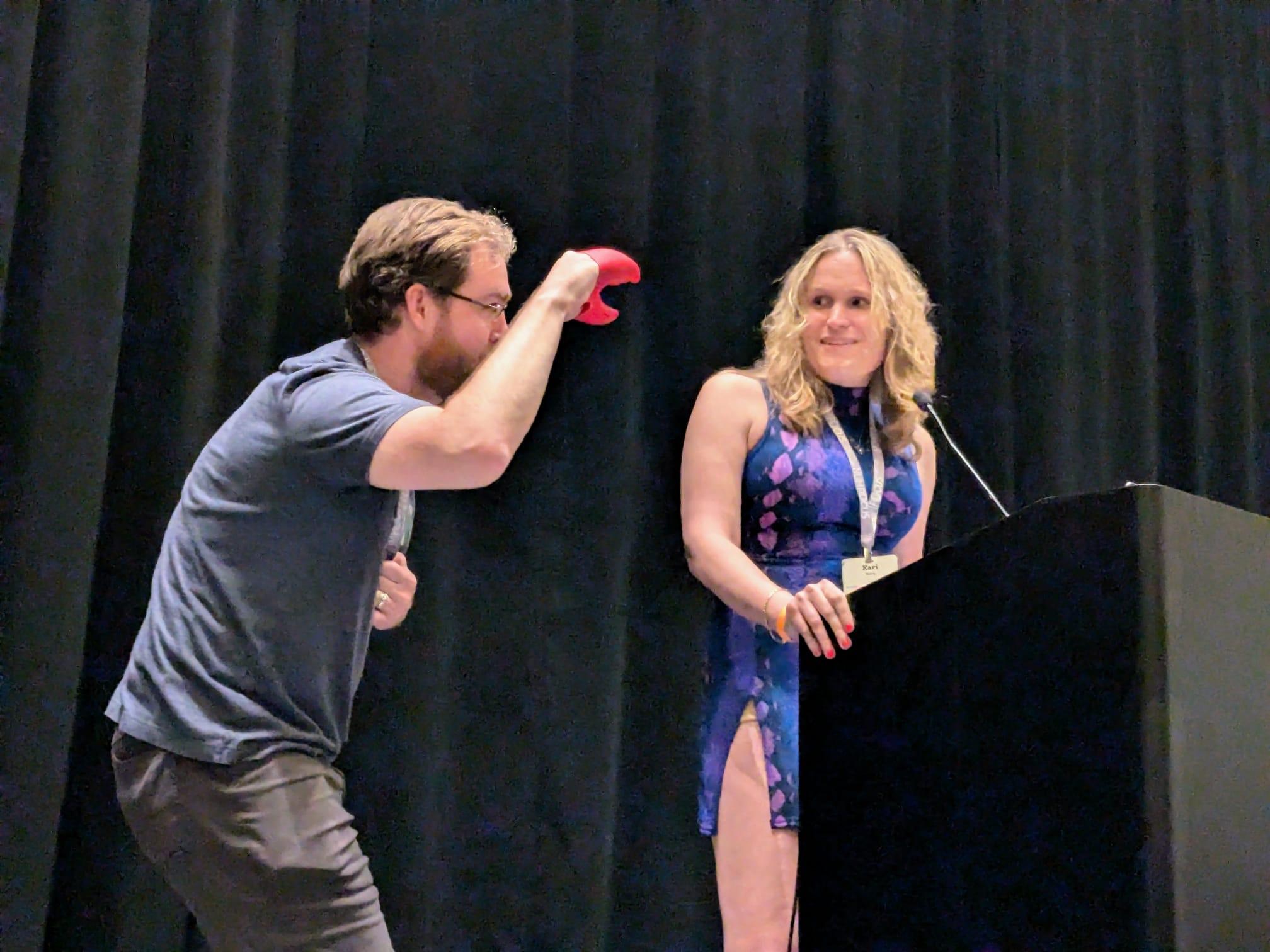 Photograph showing a man with a beard holding a red lobster claw - while deviously looking at a blonde woman at a podium who is smirking at him. She is wearing a blue top and beige pants